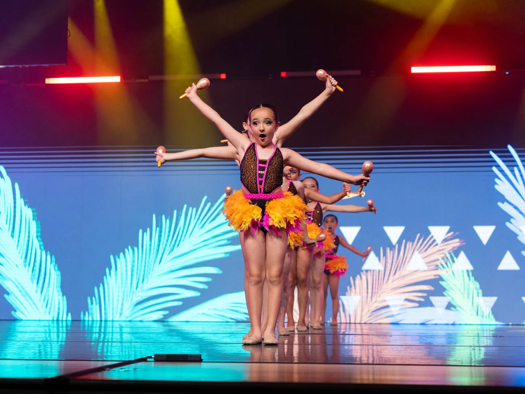 A small dancer with maracas, arms thrown wide, feathers and palm fronds behind her